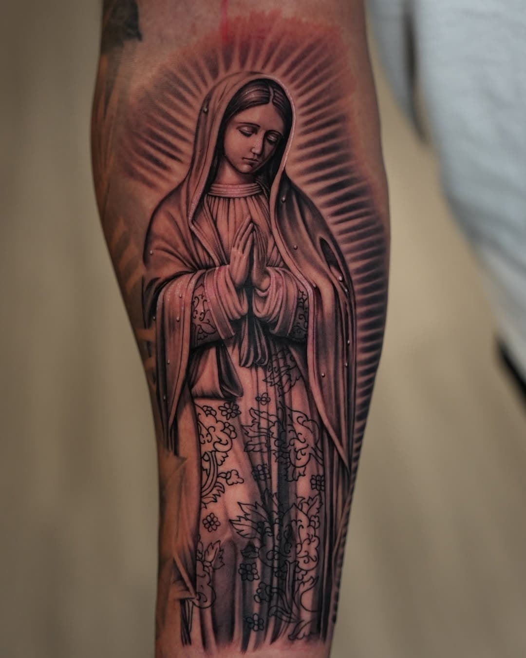 Black and grey Virgin Mary praying with radiant halo tattoo on forearm by Ink Theory
